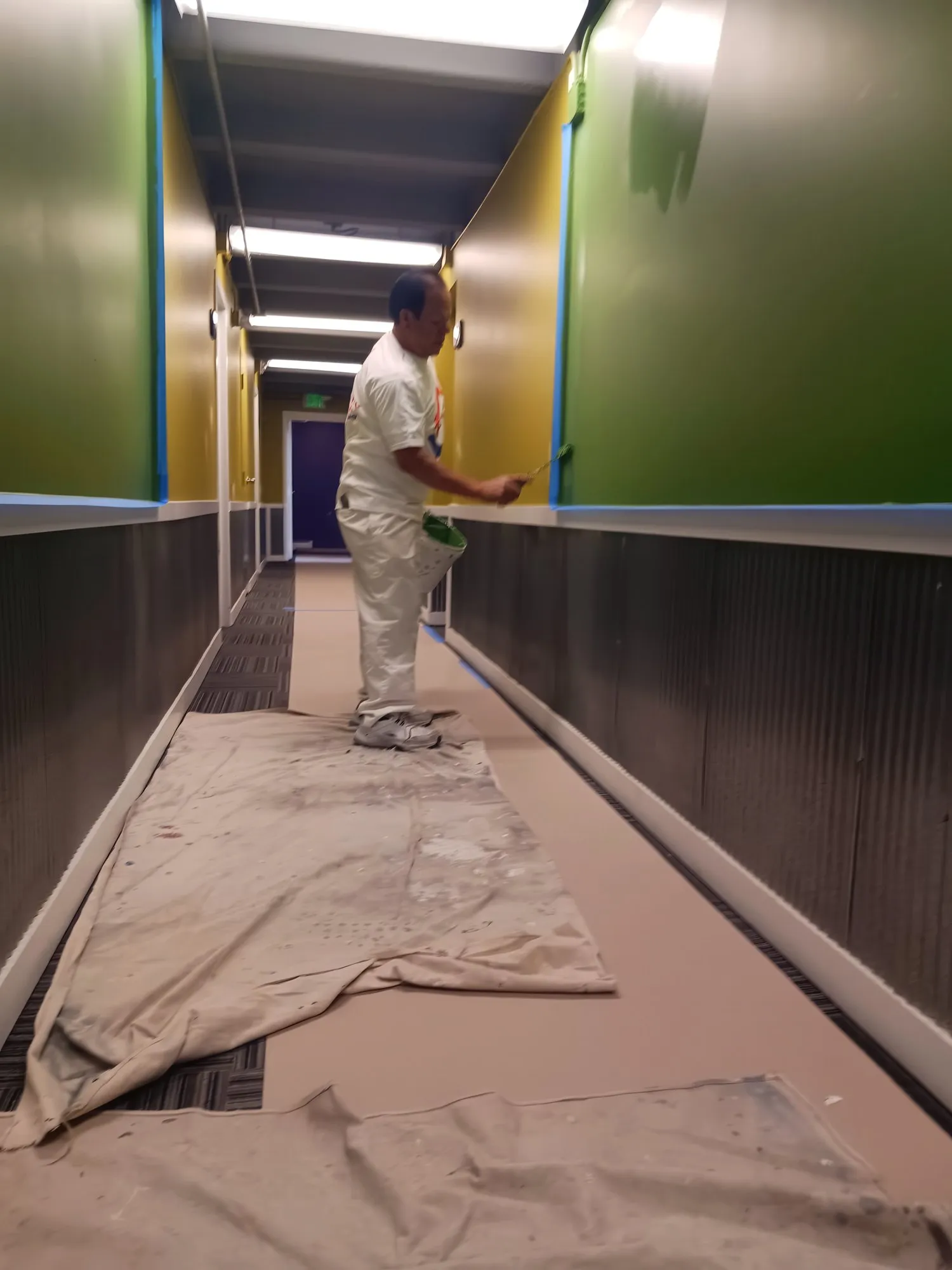 Jose painting the walls of a long commercial hallway with floors covered